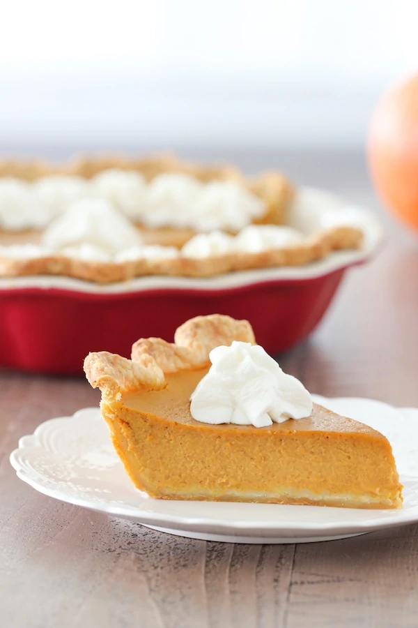A slice of smooth and tender pumpkin pie with whipped cream and a deep dish pie plate in the background