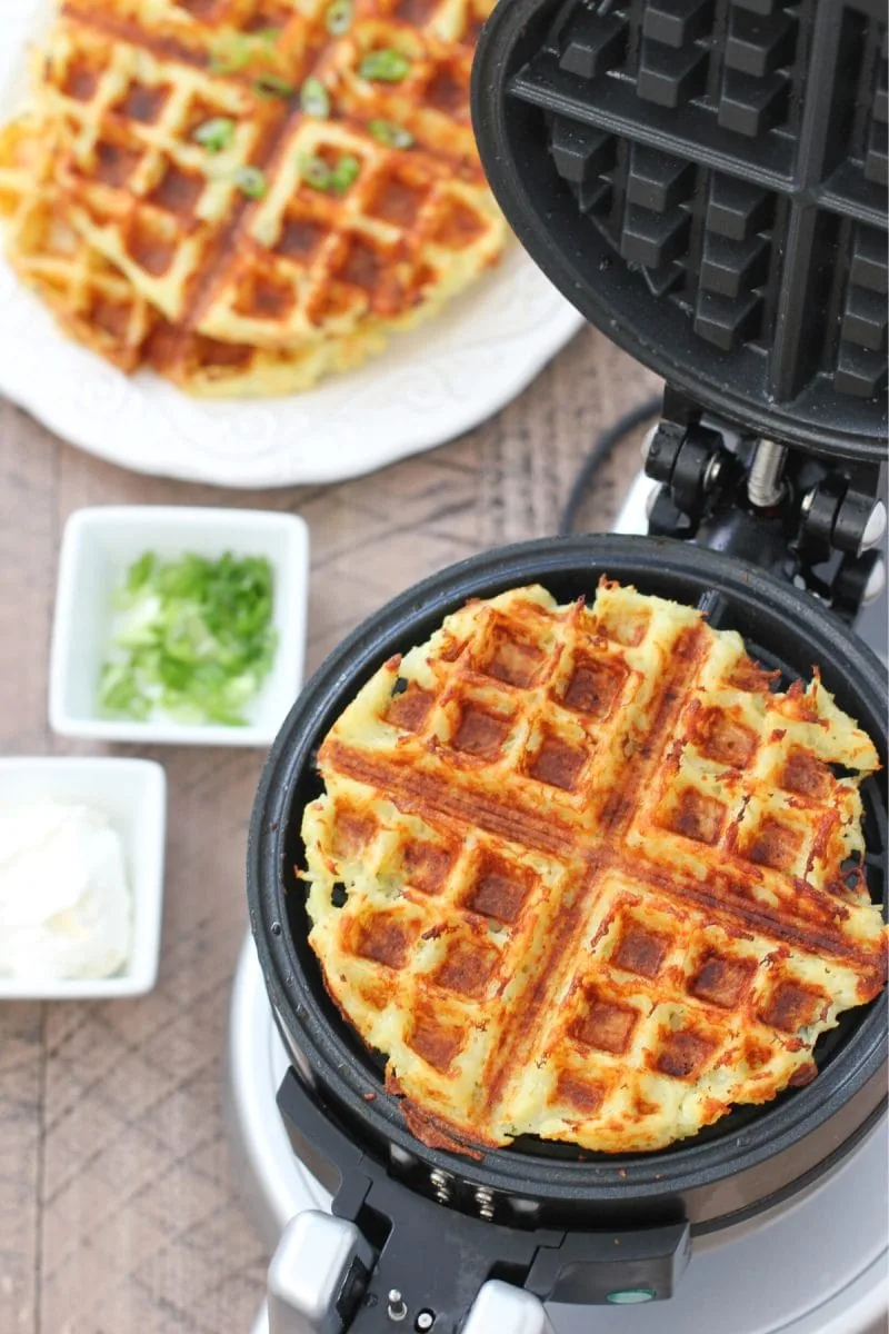 Potato waffles cooking in a waffle maker with green onions and sour cream on the side.