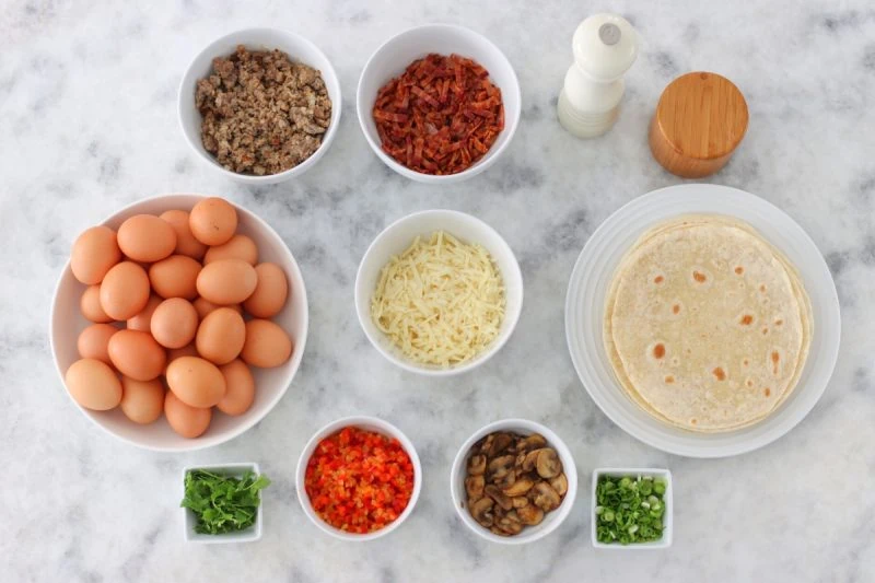 Meal Prep Breakfast Burritos Ingredients (eggs, sausage, bacon, grated cheese, fresh herbs, cilantro, green onions, bell peppers, onions, mushrooms, tortillas)