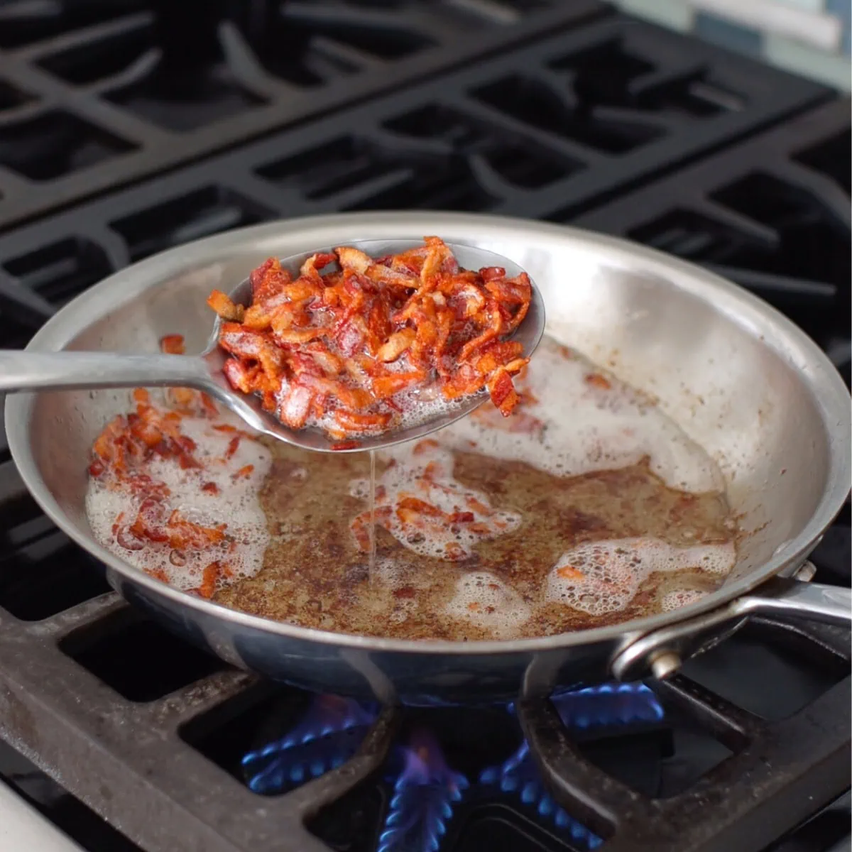 Bacon cooking in a pan, large spoon scooping out a portion. 