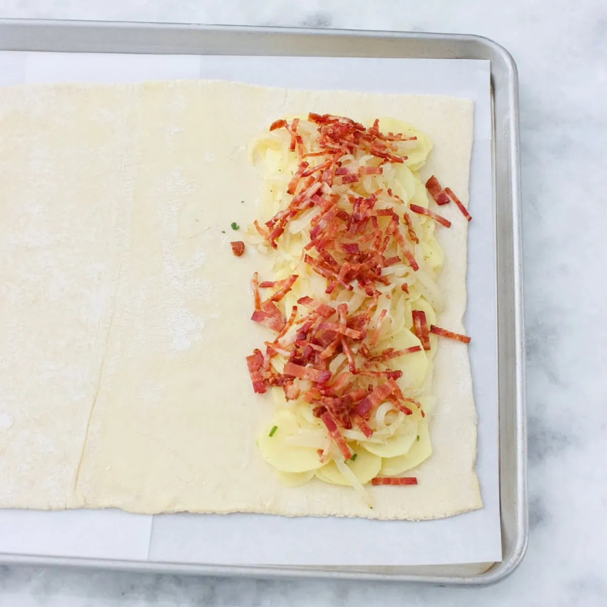 Assembling Potato bacon pastry before baking. puff pastry on a baking sheet, topped with potatoes, onions and bacon.  